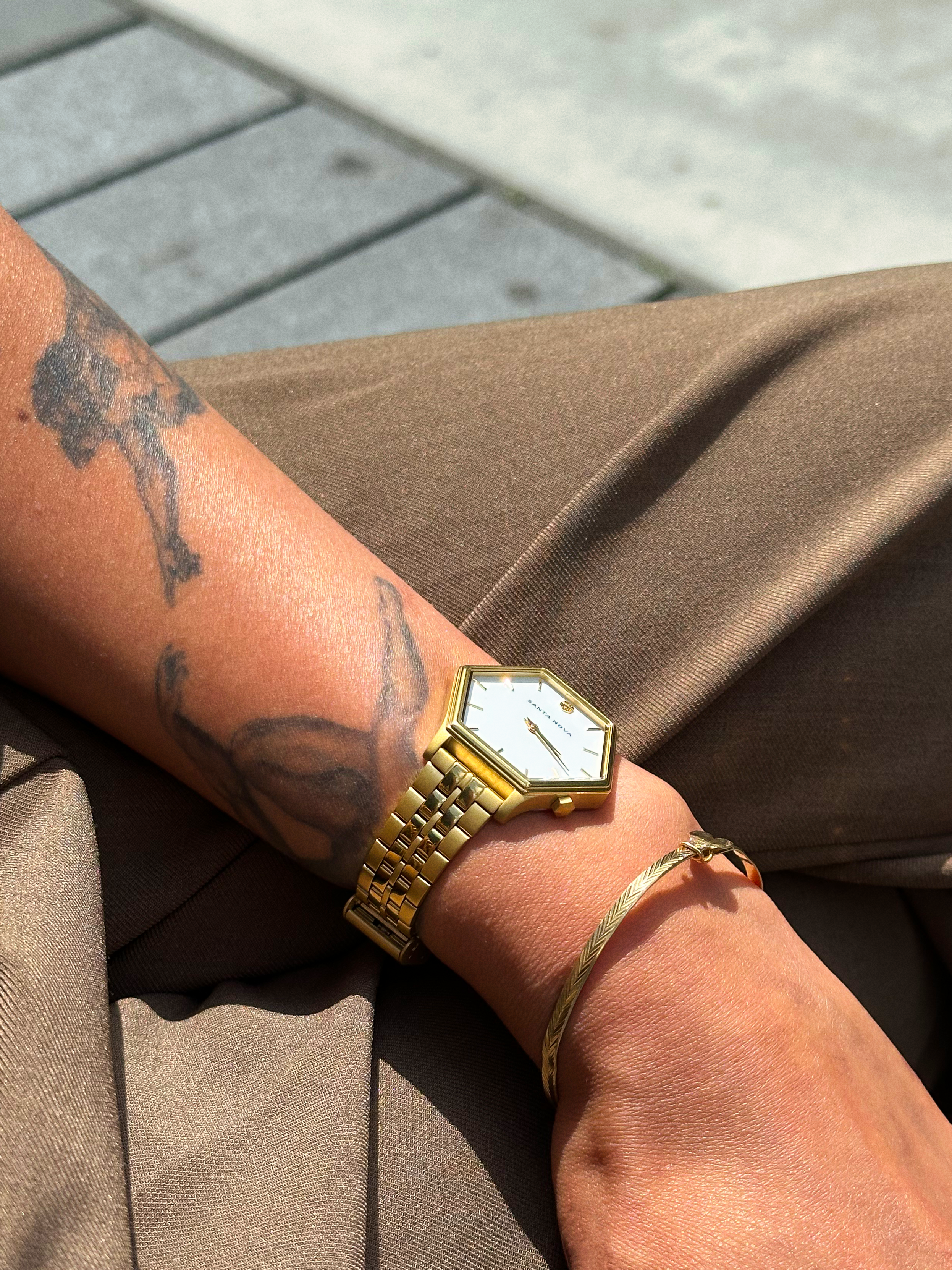 Gold wristwatch on a person's wrist with a tattooed arm, wearing brown pants.